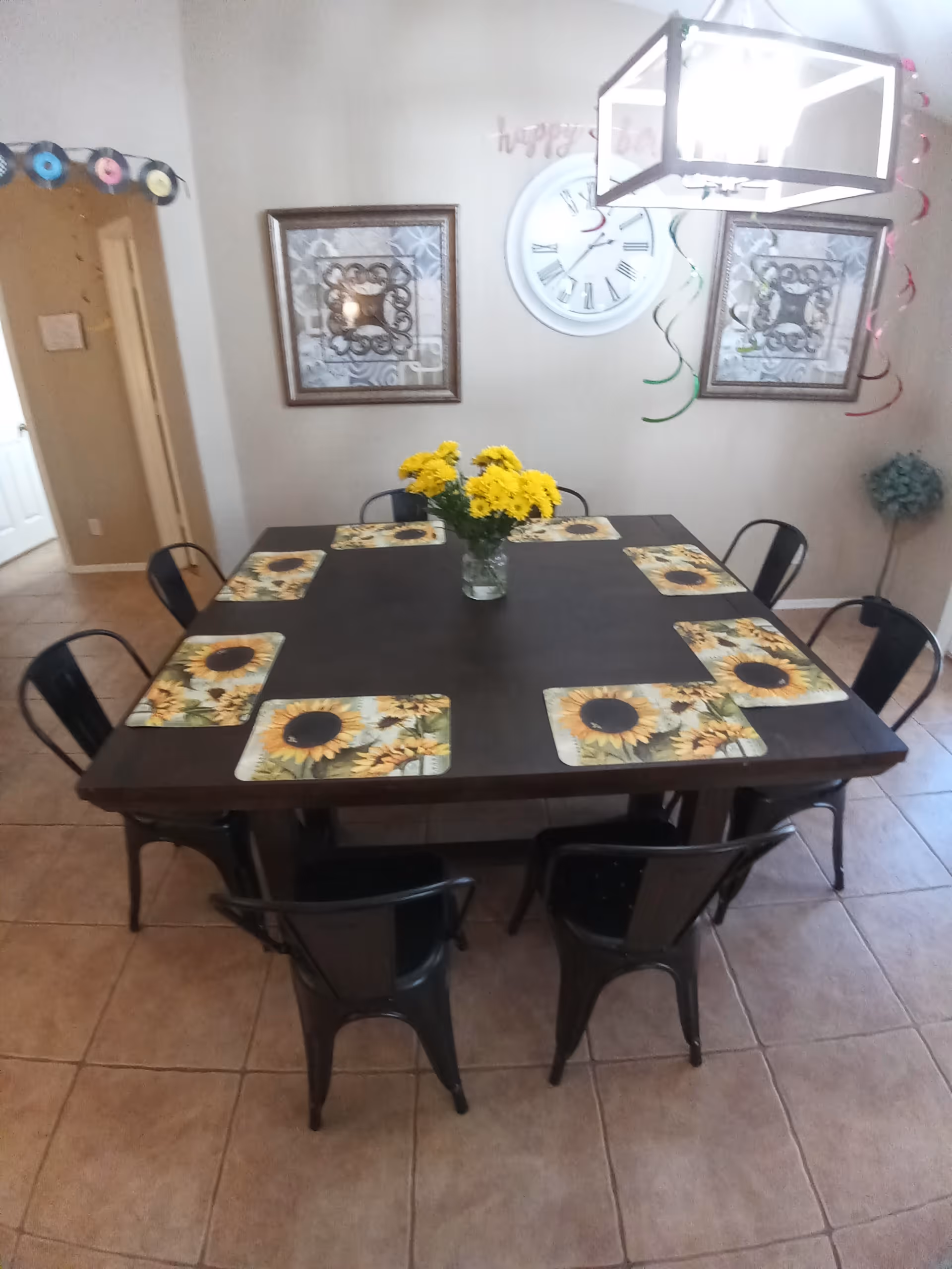 A dining room with a dark wooden square table set with eight black metal chairs. The table has sunflower-themed placemats and a vase with yellow flowers in the center. The walls have two framed decorative artworks and a large round clock. Colorful hanging streamers and a 'happy birthday' banner are visible, along with a modern rectangular ceiling light fixture.