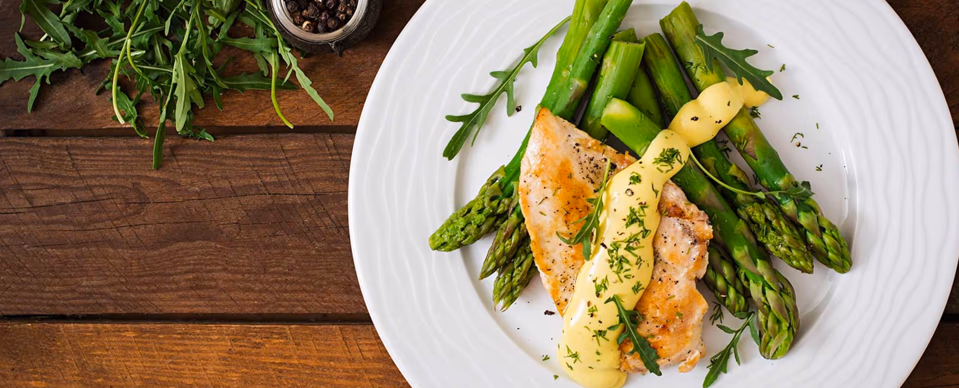 A white plate with cooked asparagus and two pieces of grilled chicken breast topped with a creamy yellow sauce and garnished with fresh herbs, placed on a wooden table with some green leafy herbs and a small container of black pepper in the background.