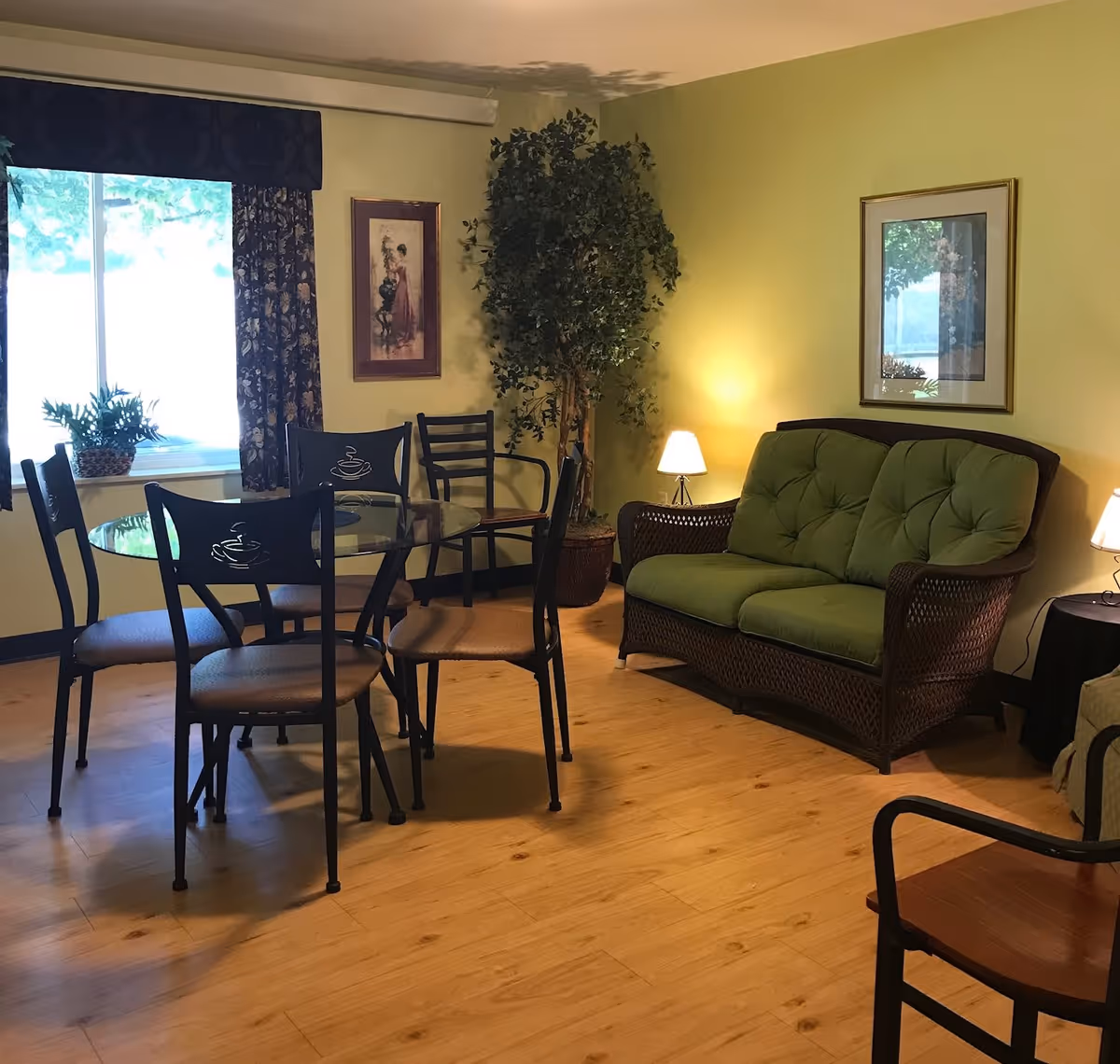 A cozy interior room with a round glass dining table surrounded by four chairs, a green cushioned wicker loveseat, two small table lamps, a large potted plant, framed artwork on the walls, and a window with floral curtains letting in natural light.