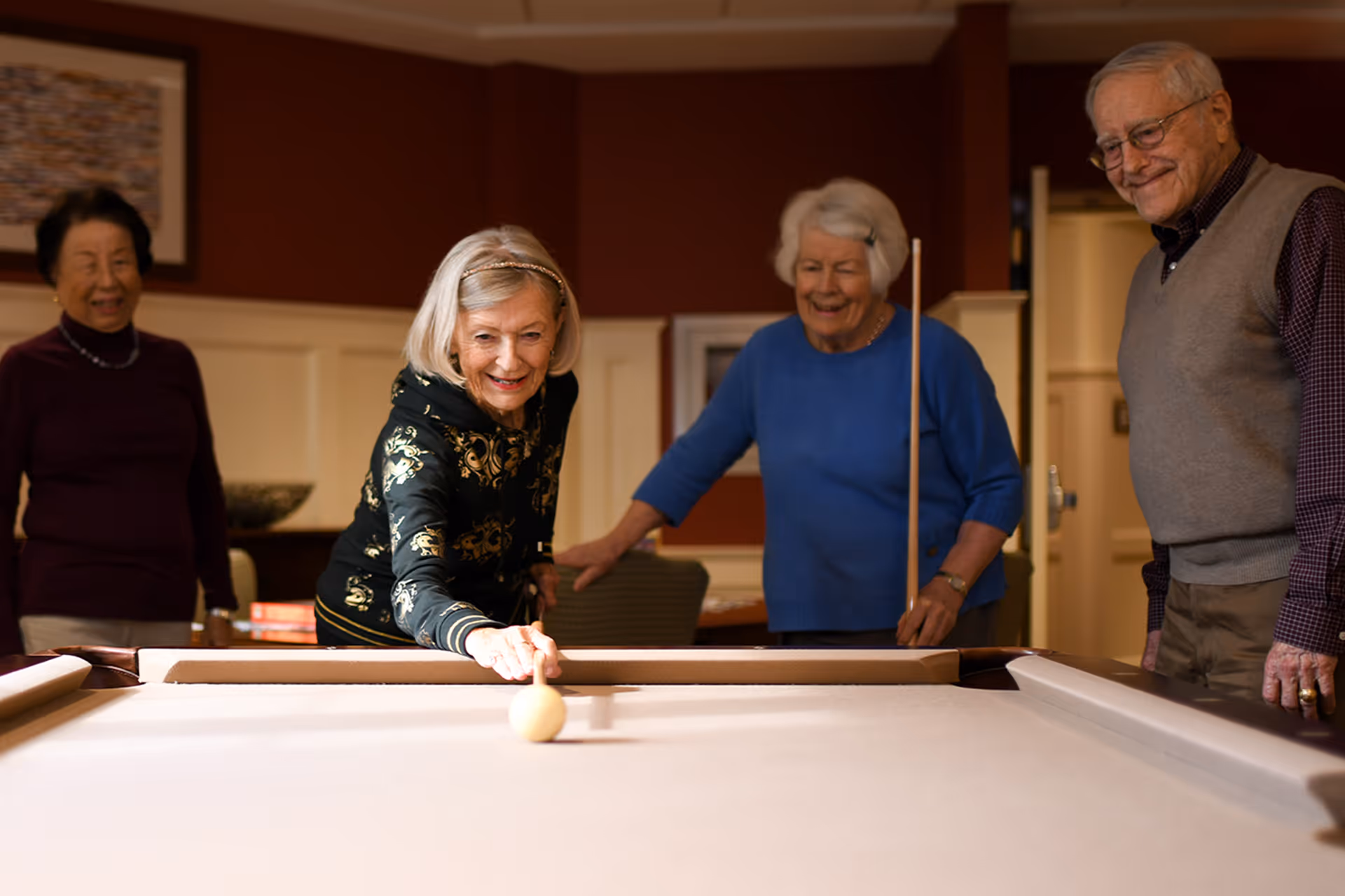 Four elderly people playing pool together in a warmly lit room. One woman is taking a shot while the others watch and smile. The room has red walls and white wainscoting with framed artwork in the background.