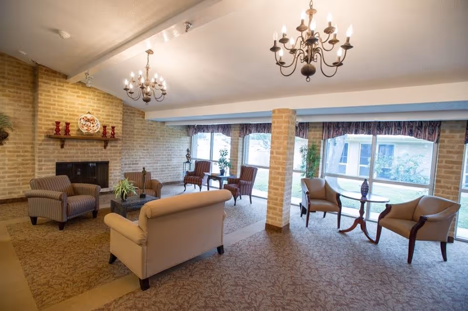 Bright communal lounge with sofas and armchairs arranged around a fireplace under chandeliers, with large windows overlooking a courtyard.
