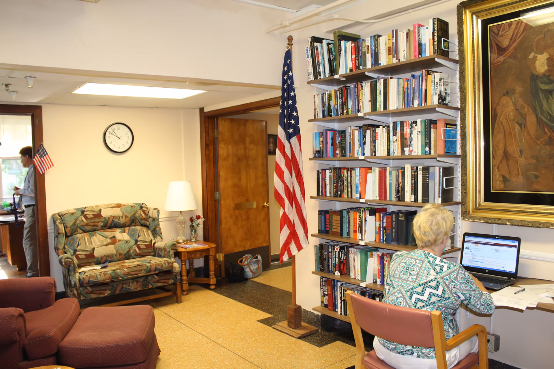 A cozy room with a woman sitting at a desk using a laptop. Behind her is a tall bookshelf filled with books and an American flag on a stand. To the left, there is a patterned loveseat, a small wooden side table with a lamp and flowers, and a clock on the wall. A man is visible in an adjacent room through an open doorway.