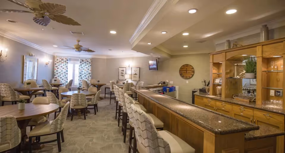 A well-lit dining area with multiple round tables and cushioned chairs arranged neatly. There is a long granite countertop bar with high chairs on one side and wooden cabinetry with glass shelves displaying decorative items on the other. The room has ceiling fans, wall sconces, framed artwork, and a wall-mounted TV. Light-colored walls and patterned curtains cover the windows at the far end.