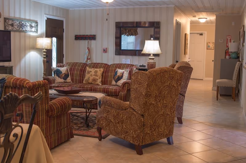A cozy living room with patterned armchairs and a striped sofa adorned with rooster-themed pillows. There is a wooden coffee table on a rug, a table lamp on a side table, a large mirror on the wall, and a hallway leading to other rooms. The room has tiled floors and paneled walls.