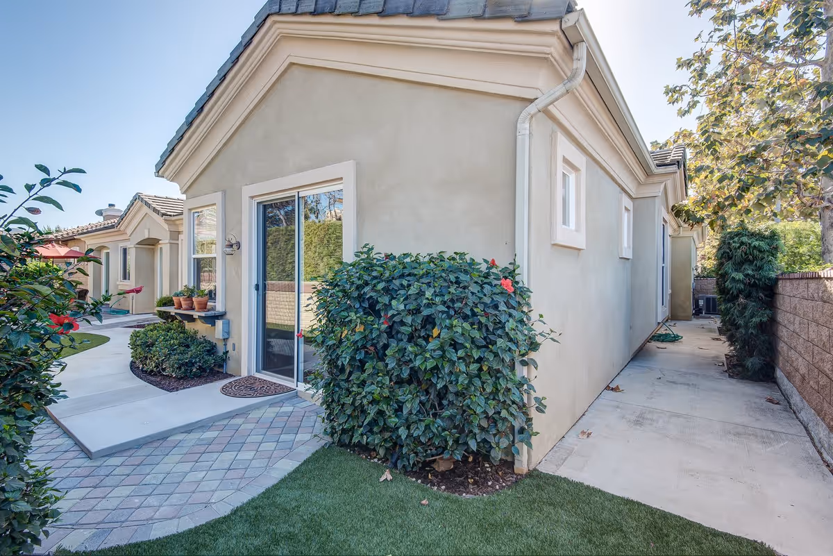 Exterior view of a beige stucco building with a tiled roof, featuring a sliding glass door, potted plants on a small shelf, and a well-maintained garden with bushes and a paved walkway under a clear blue sky.