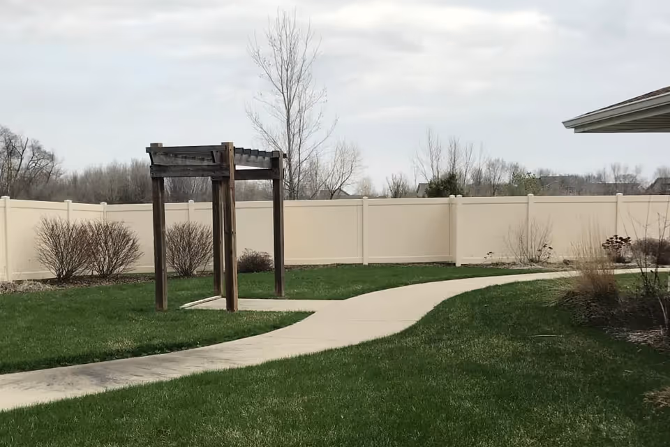 A curved concrete pathway runs through a green lawn with a wooden pergola structure on the left side. There are leafless bushes and trees along a beige privacy fence in the background under a cloudy sky.