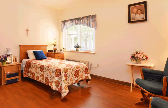 A cozy bedroom with a single bed covered in a floral bedspread, a wooden nightstand with a lamp and flowers, a window with blue curtains, a wooden chair with blue cushions, a small table with a flower arrangement, a cross on the wall, and a framed photo of an elderly couple.