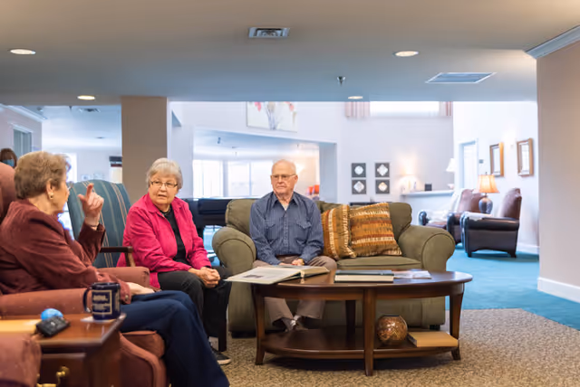 Three elderly residents sit in a comfortable communal lounge area with sofas and a coffee table.