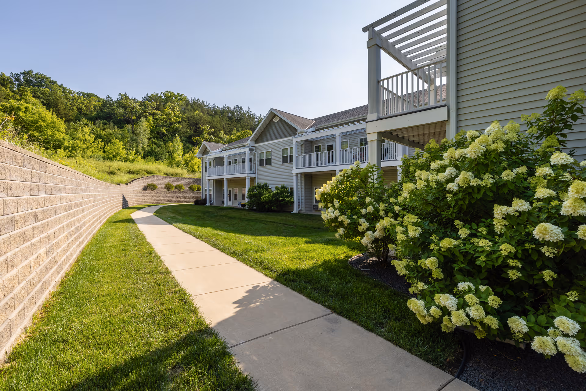 Sidewalk leading alongside a two-story senior living building with balconies, landscaped lawn and flowering shrubs under a clear sky.