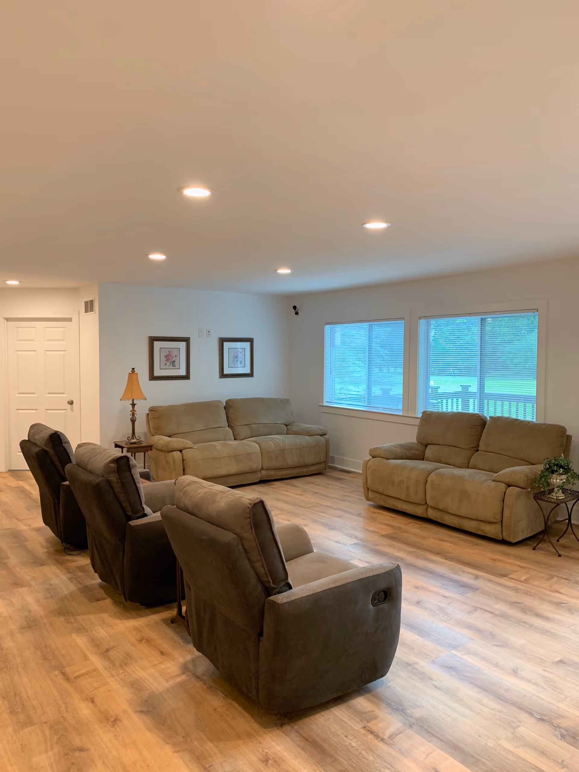 A spacious living room with wooden flooring, two beige sofas against the walls, three dark brown recliner chairs in the center, a small side table with a lamp, two framed pictures on the wall, and two large windows with blinds letting in natural light.