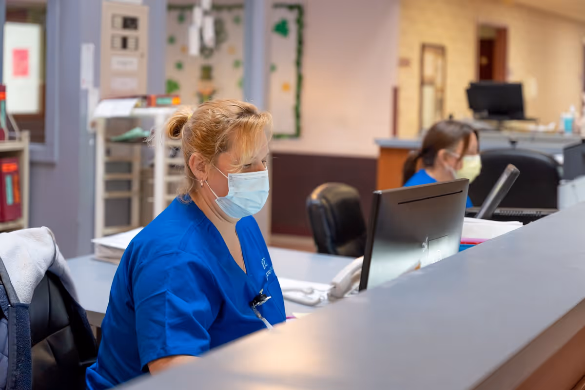 Two healthcare workers wearing blue scrubs and face masks sitting at a reception desk working on computers in a care facility.