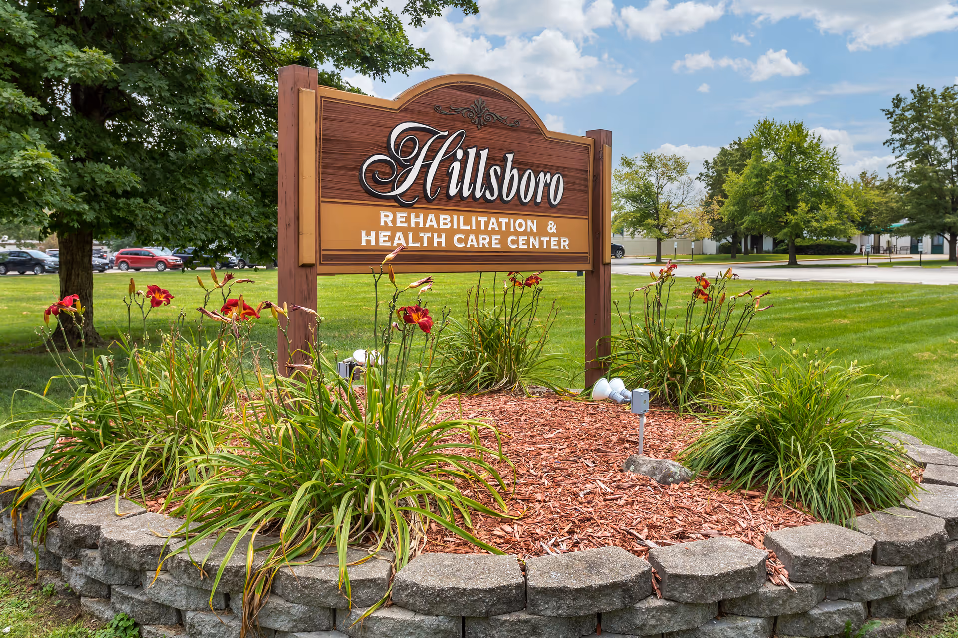 A wooden sign for Hillsboro Rehabilitation & Health Care Center surrounded by a circular flower bed with green plants and red flowers, set on a grassy lawn with trees and a partly cloudy sky in the background.