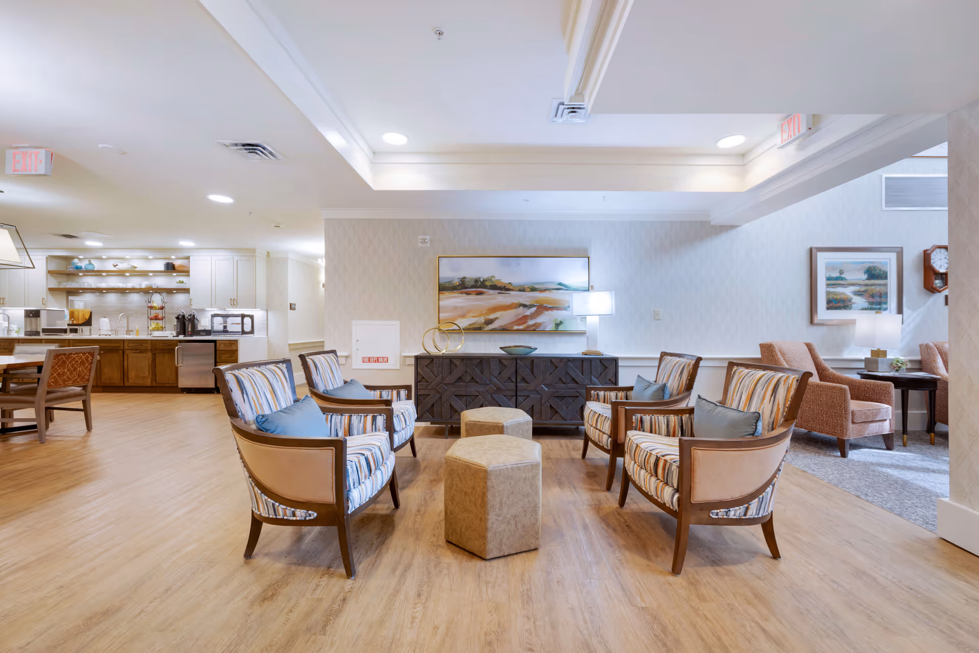 A bright and spacious common area in a senior living facility featuring four striped armchairs with blue cushions arranged around two hexagonal ottomans on a wood floor. In the background, there is a dark wooden sideboard with a lamp, decorative rings, and a landscape painting above it. To the left, there is a kitchen area with wooden cabinets, open shelves with dishes, and a dining table with chairs. To the right, there is a seating area with two orange armchairs, a side table with a lamp, a wall clock, and a framed painting.