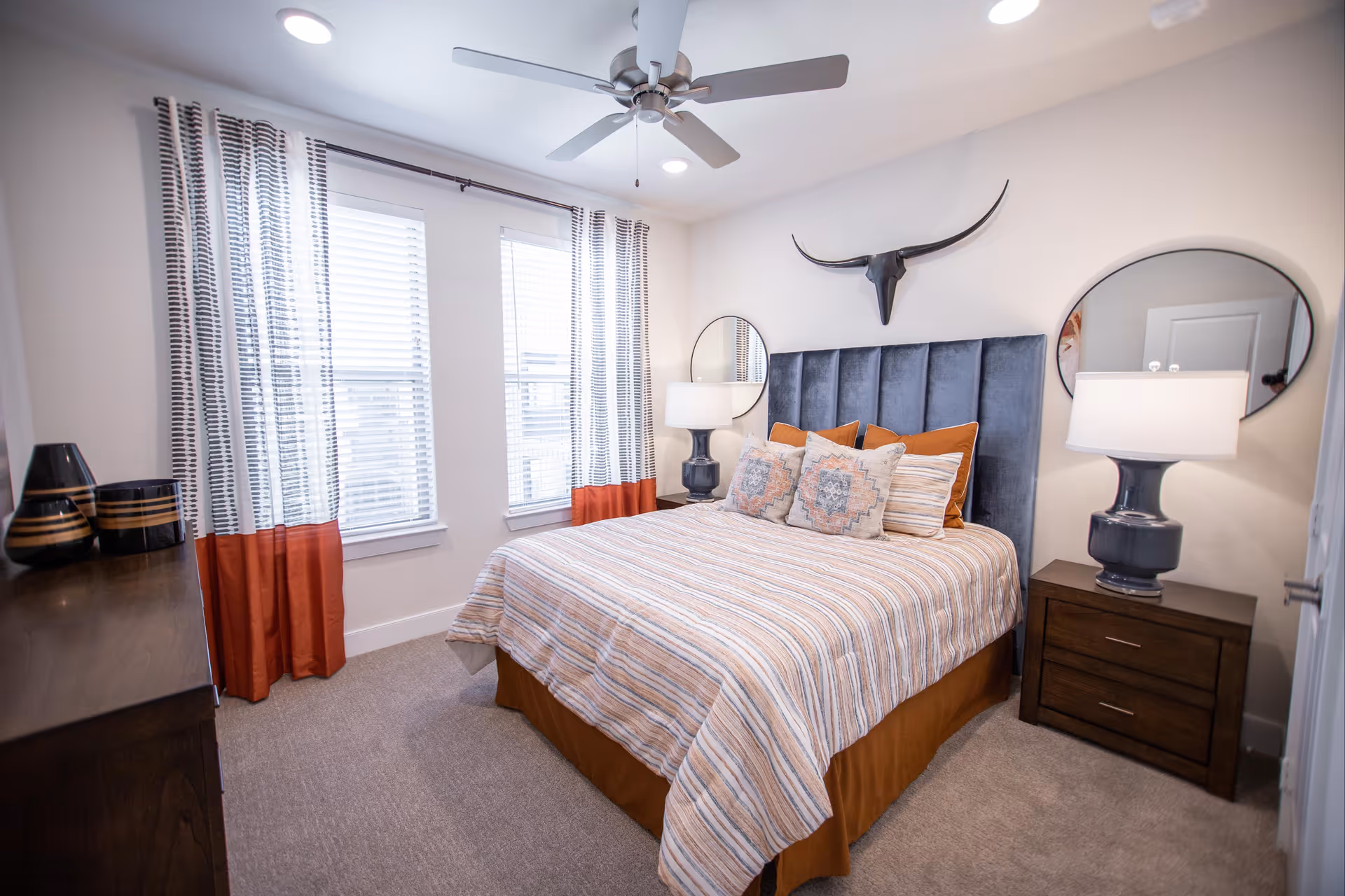 A neatly arranged bedroom with a bed featuring a striped bedspread and multiple pillows. The bed has a tall, dark blue upholstered headboard. On either side of the bed are dark wooden nightstands with matching lamps. Above the headboard is a decorative longhorn skull mounted on the wall. Two round mirrors hang on the wall behind each nightstand. The room has two windows with white blinds and curtains that are white with black patterns and orange at the bottom. A ceiling fan with lights is mounted on the ceiling.