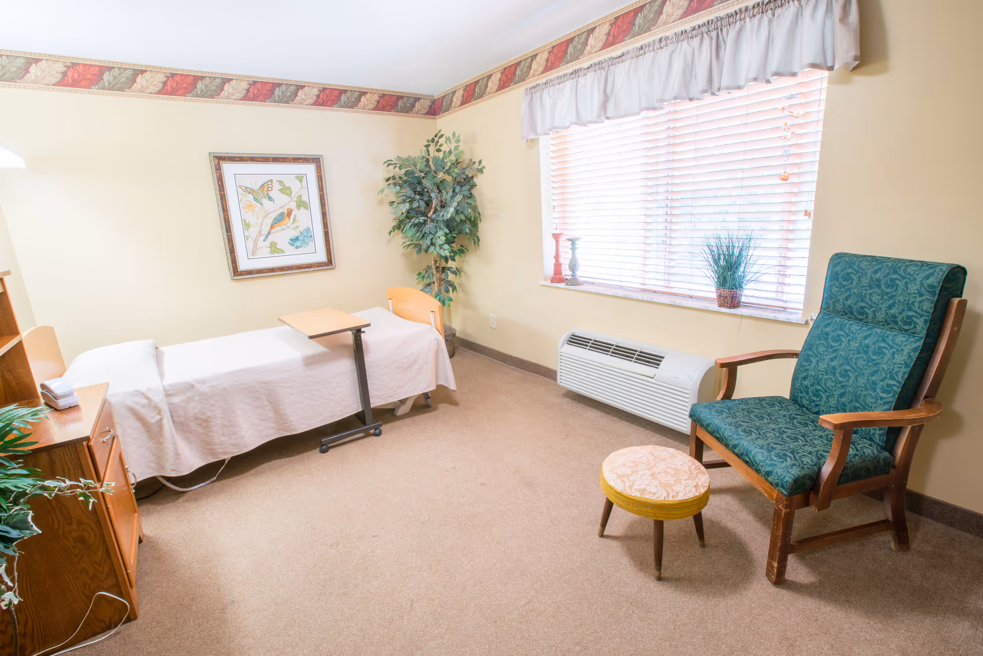 A small, cozy room with a single bed covered in a white blanket, a wooden bedside table with a lamp, a framed picture of birds on the wall, a potted plant in the corner, a large window with blinds and a valance, a green upholstered armchair with wooden arms, and a small round footstool.