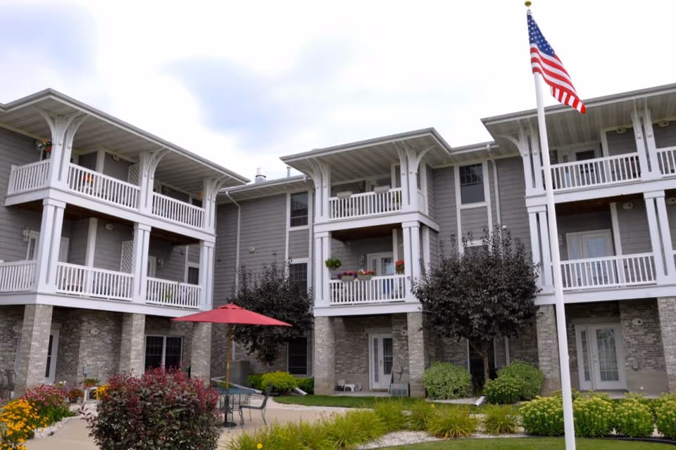 Three-story gray and white senior living building with multiple balconies, a landscaped courtyard with seating and an American flag.
