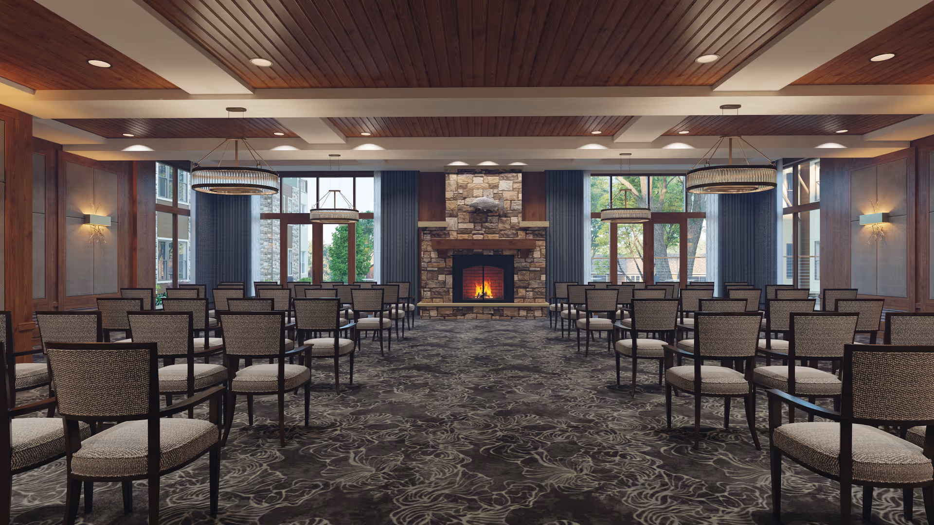 A spacious room with rows of chairs arranged facing a stone fireplace with a fire burning. The room features large windows with blue curtains, wooden ceiling beams, and modern circular chandeliers. The carpet has a floral pattern in dark tones.