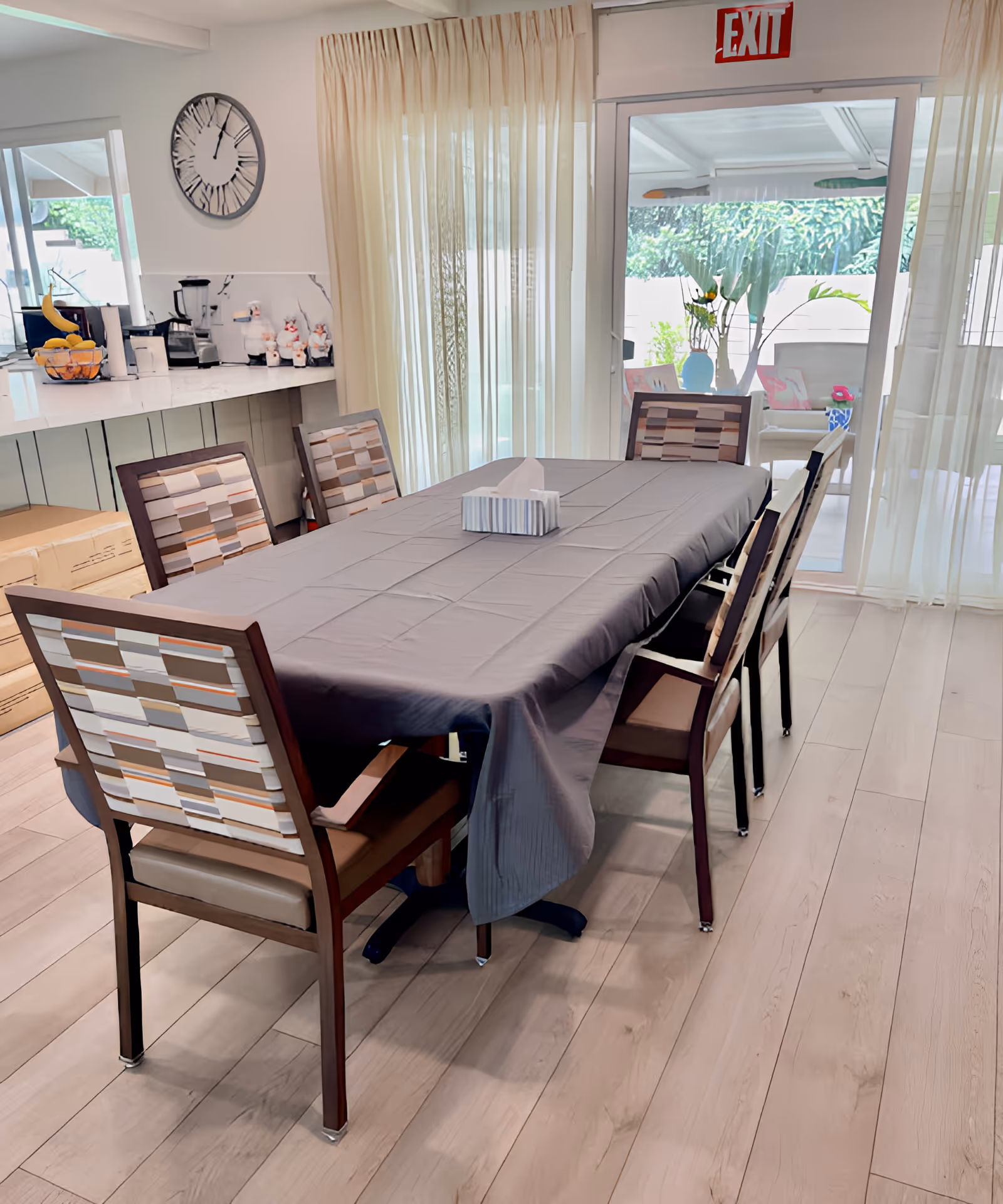 Dining area with a rectangular table covered with a gray tablecloth and six chairs with patterned backs. Behind the table is a counter with kitchen appliances and a large clock on the wall. Sheer curtains cover a sliding glass door leading to an outdoor patio with plants and seating. An exit sign is visible above the door.