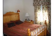 A bedroom with a wooden bed frame and headboard, a bed covered with a red and brown striped bedspread and two dark pillows. Next to the bed is a wooden nightstand with a white lamp and some small items. The window is covered with floral patterned curtains.