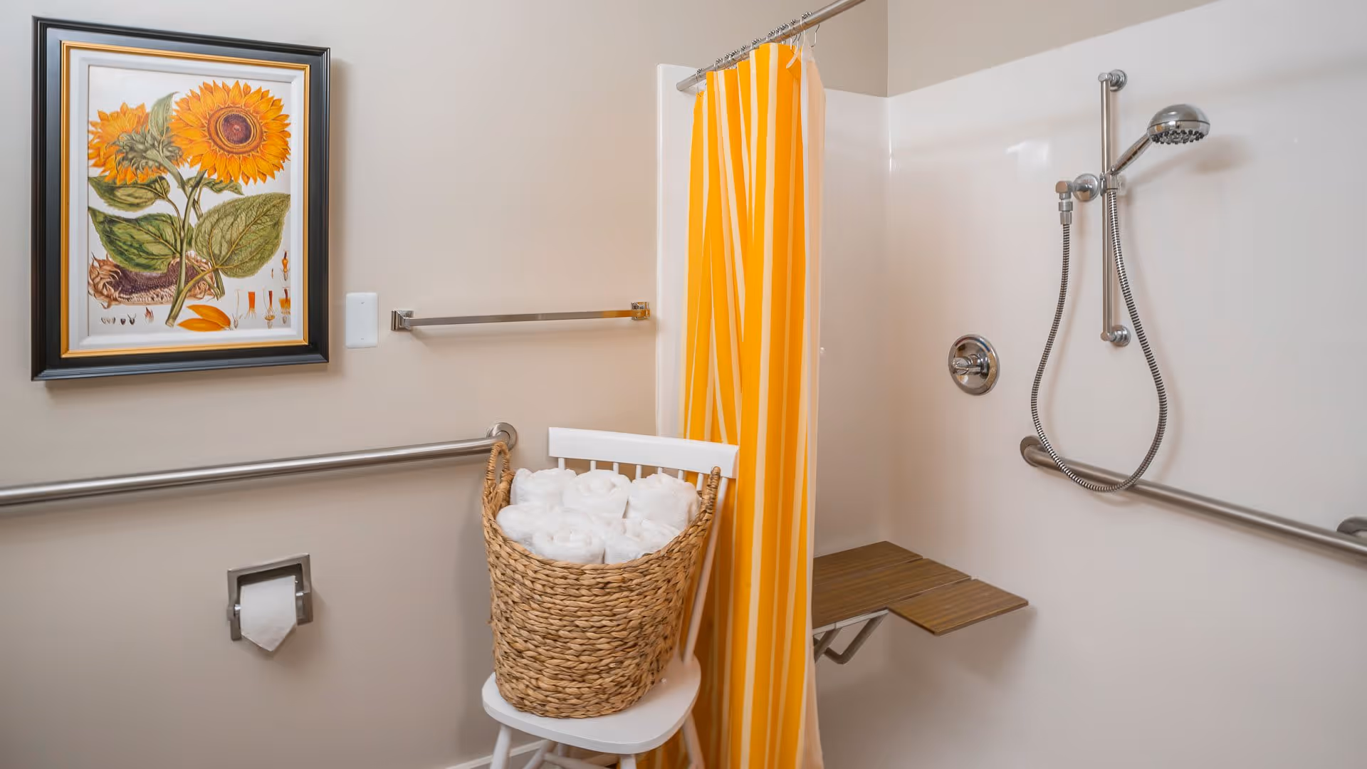 Bathroom with a walk-in shower featuring a handheld showerhead and a fold-down wooden seat. A yellow and white striped shower curtain is partially drawn. There are grab bars installed on the walls for safety. A white chair holds a wicker basket filled with rolled white towels. A framed sunflower artwork hangs on the wall above a toilet paper holder.