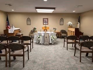 A small chapel or prayer room with rows of chairs facing an altar decorated with flowers and religious items. The room has beige walls, a carpeted floor, and an American flag on the left side. A framed picture hangs on the wall behind the altar.