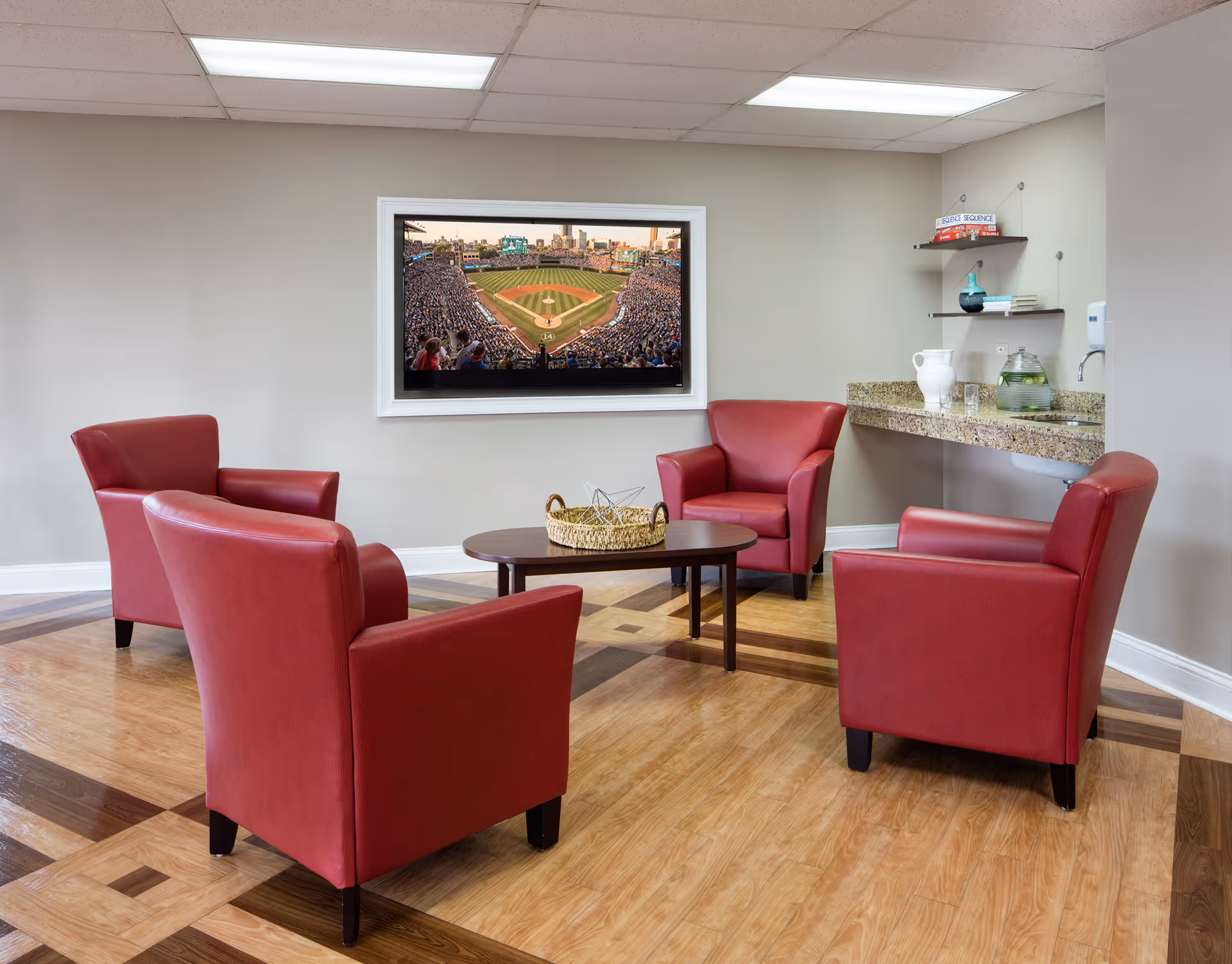 A cozy seating area with four red armchairs arranged around a wooden coffee table with a woven basket on top. A large flat-screen TV mounted on the wall displays a baseball stadium filled with spectators. To the right, there is a granite countertop with a pitcher, glasses, and decorative items on shelves above. The room has a patterned wooden floor and neutral-colored walls.