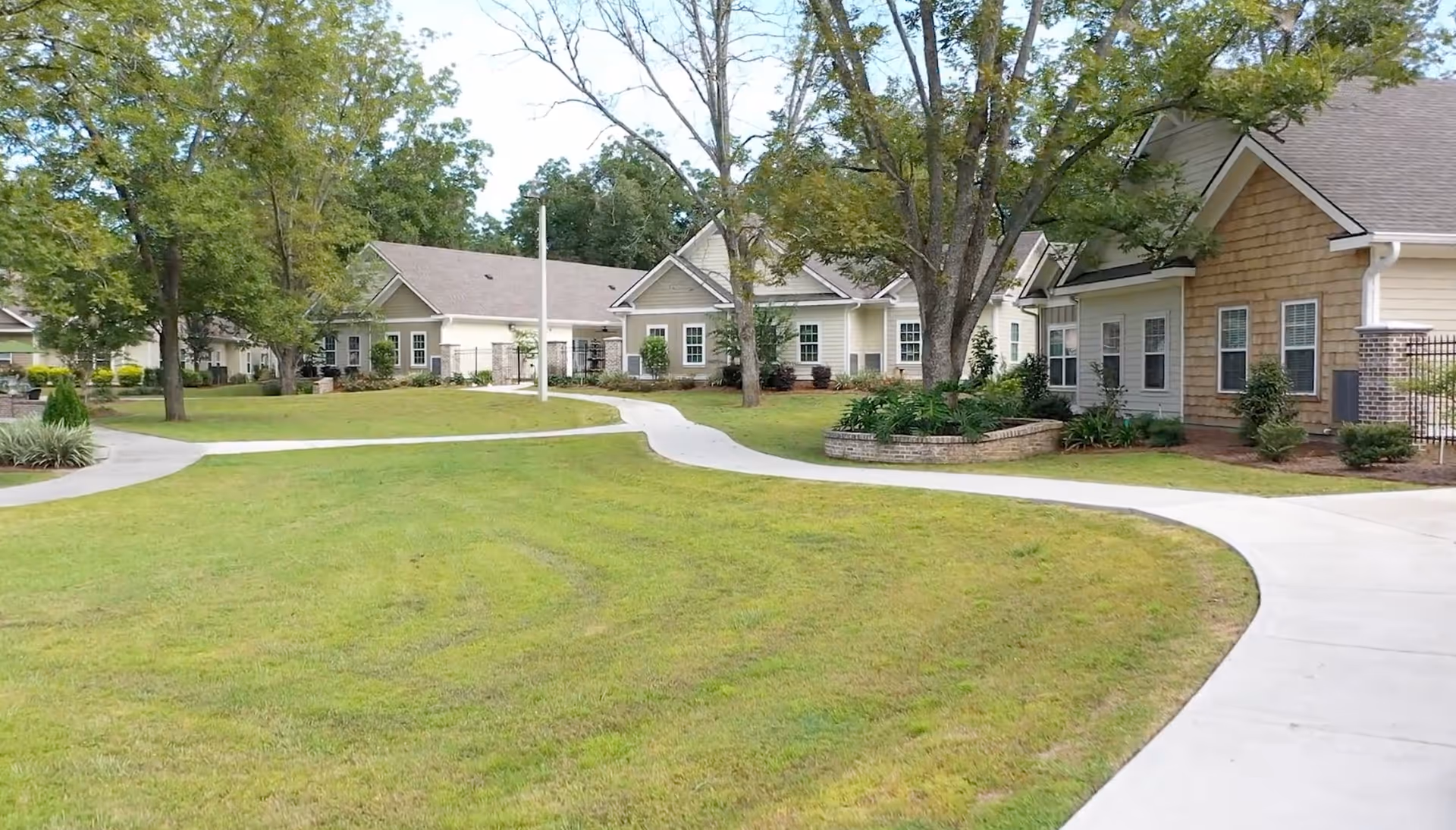 Outdoor view of a senior living facility with multiple single-story buildings surrounded by green lawns, trees, and paved walkways.