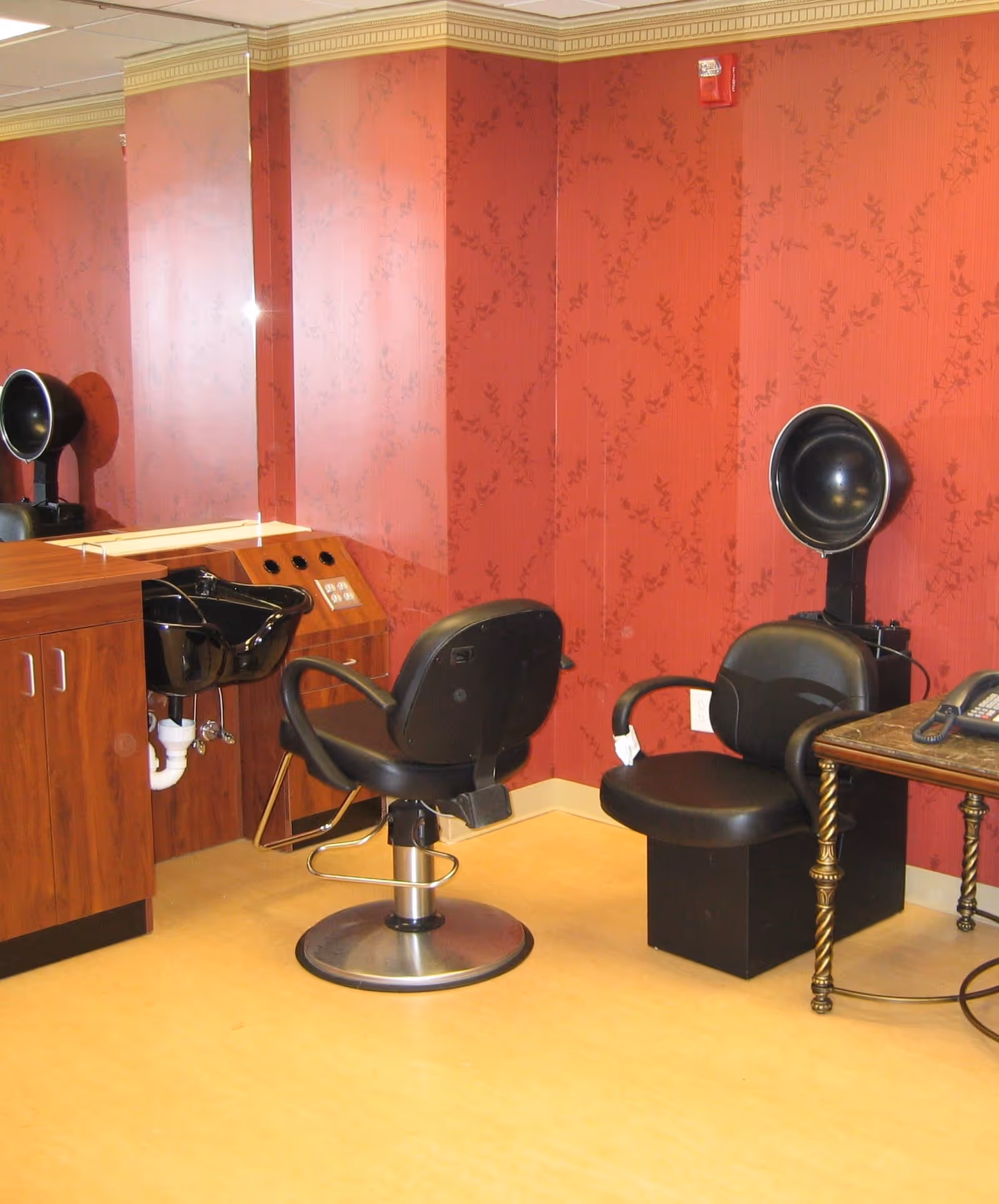Interior of a salon area with a black salon chair in front of a black sink, a black hair dryer chair with a hooded dryer, a wooden cabinet, and a small table with a telephone. The walls are red with a subtle floral pattern.