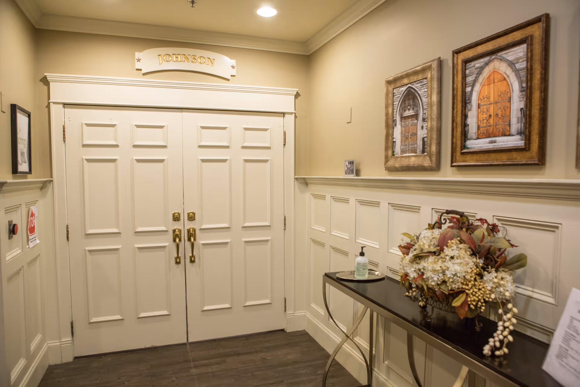A hallway with double white paneled doors labeled 'Johnson' above. The walls are beige with white wainscoting. On the right side, there is a black console table with a floral arrangement and a bottle of hand sanitizer. Above the table, two framed pictures of arched wooden doors hang on the wall.