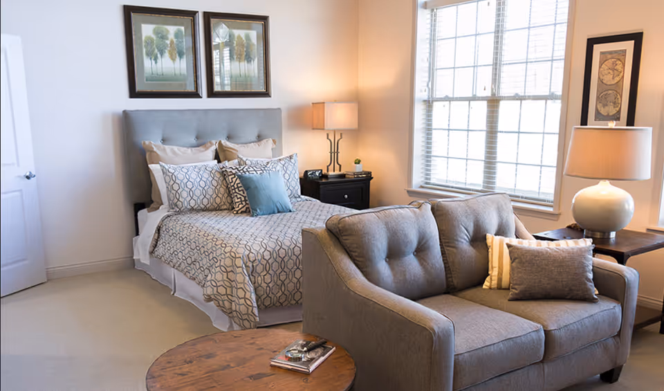 A well-lit bedroom featuring a neatly made bed with patterned bedding and multiple pillows. There is a gray upholstered headboard, two framed pictures of trees above the bed, and a nightstand with a lamp and small decor items. In front of the bed is a gray cushioned loveseat with two pillows, and next to it is a wooden side table with a large lamp. A round wooden coffee table with a book and glasses is in the foreground. Large windows with blinds allow natural light into the room.
