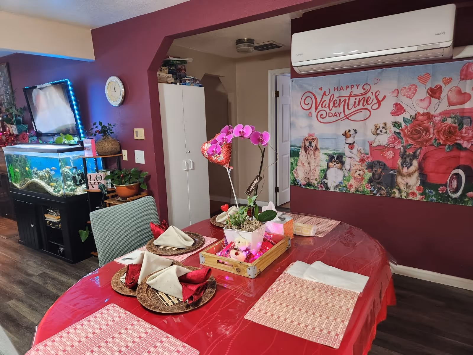 Dining area with a red tablecloth and four place settings, a pink orchid centerpiece, an aquarium, and a 'Happy Valentine's Day' wall banner.