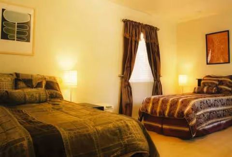 Two side-by-side beds with matching bedding, lamps, and a window between them in a lit bedroom.