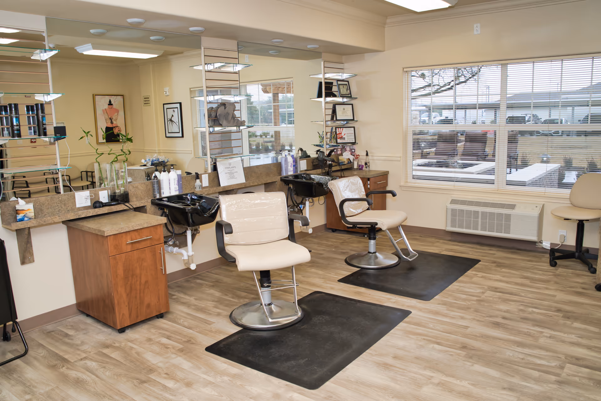 Salon area in a senior living facility with two styling chairs and wash basins in front of large mirrors and windows.