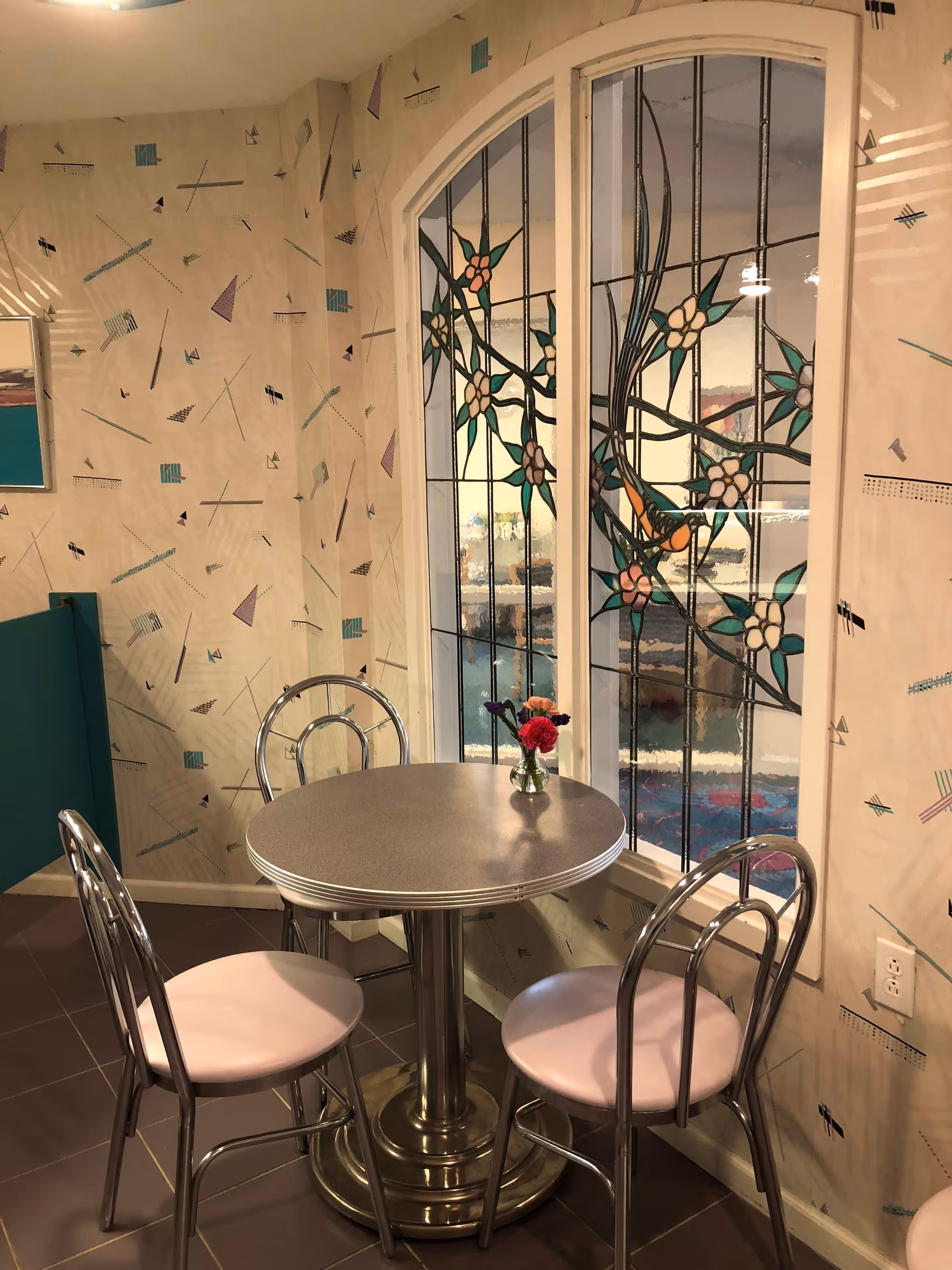 A small round table with a metallic base and three metal chairs with light pink cushions arranged around it. On the table is a small vase with colorful flowers. Behind the table is a large window with decorative stained glass featuring floral designs. The walls have a patterned wallpaper with abstract geometric shapes in pastel colors.