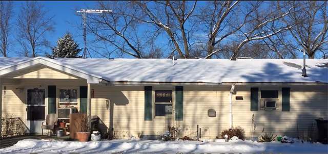 Single-story building with light yellow siding and green shutters, covered with a layer of snow on the roof and ground. There are two chairs and some potted plants near the entrance on the left side. Leafless trees are visible in the background under a clear blue sky.