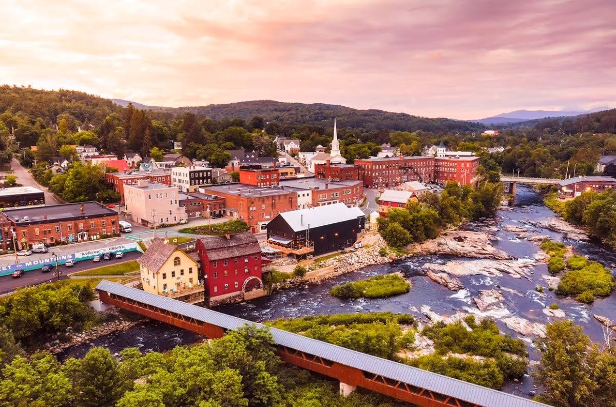A scenic aerial view of a small town with a river running through it, surrounded by lush greenery and hills under a pinkish sky at sunset. The town features a mix of historic and modern buildings, including a church with a tall steeple and a covered wooden bridge crossing the river.