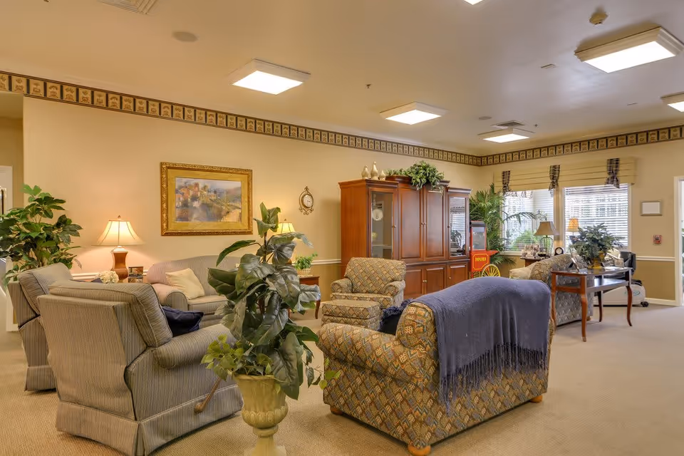 A cozy senior living common room with upholstered sofas and armchairs, plants, lamps, and a wooden cabinet under bright ceiling lights.