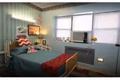 A small, neatly arranged bedroom with a single bed covered in a teal blanket and a red throw. The bed has a wooden headboard and footboard. There is a bedside table with a lamp, a small American flag, and some framed pictures on the wall. A window air conditioning unit is installed in the window, which has blinds partially drawn. The walls are decorated with light green striped wallpaper and a floral border near the ceiling.
