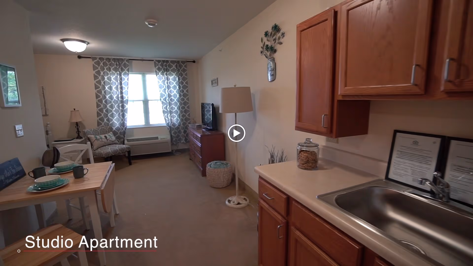 Interior view of a studio apartment in a senior living facility showing a small kitchenette with wooden cabinets and a sink on the right, a dining table set with plates and cups on the left, and a living area with a chair, lamp, TV, and window with patterned curtains in the background.