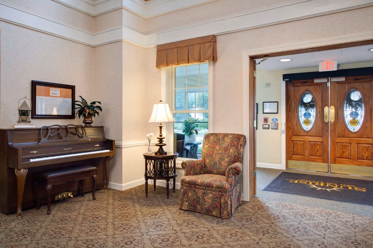 A cozy interior corner of a senior living facility featuring a wooden piano with a bench, a small round table with a lamp and a flower vase, a patterned armchair, and a window with a brown valance. In the background, double wooden doors with oval glass windows and an exit sign above are visible.