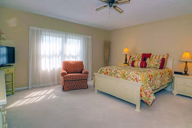 Bright bedroom with a floral bedspread, upholstered armchair, nightstands, TV and a large window with sheer curtains.