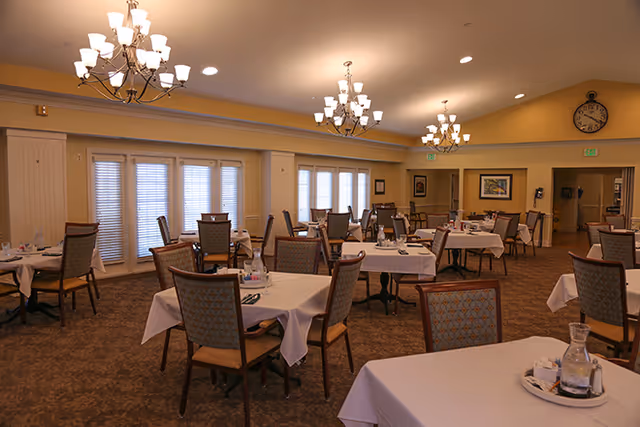 A spacious dining room with multiple tables covered in white tablecloths, each set with chairs, glass water pitchers, and napkins. The room features warm lighting from chandeliers hanging from the ceiling, large windows with blinds, and framed artwork on the walls.