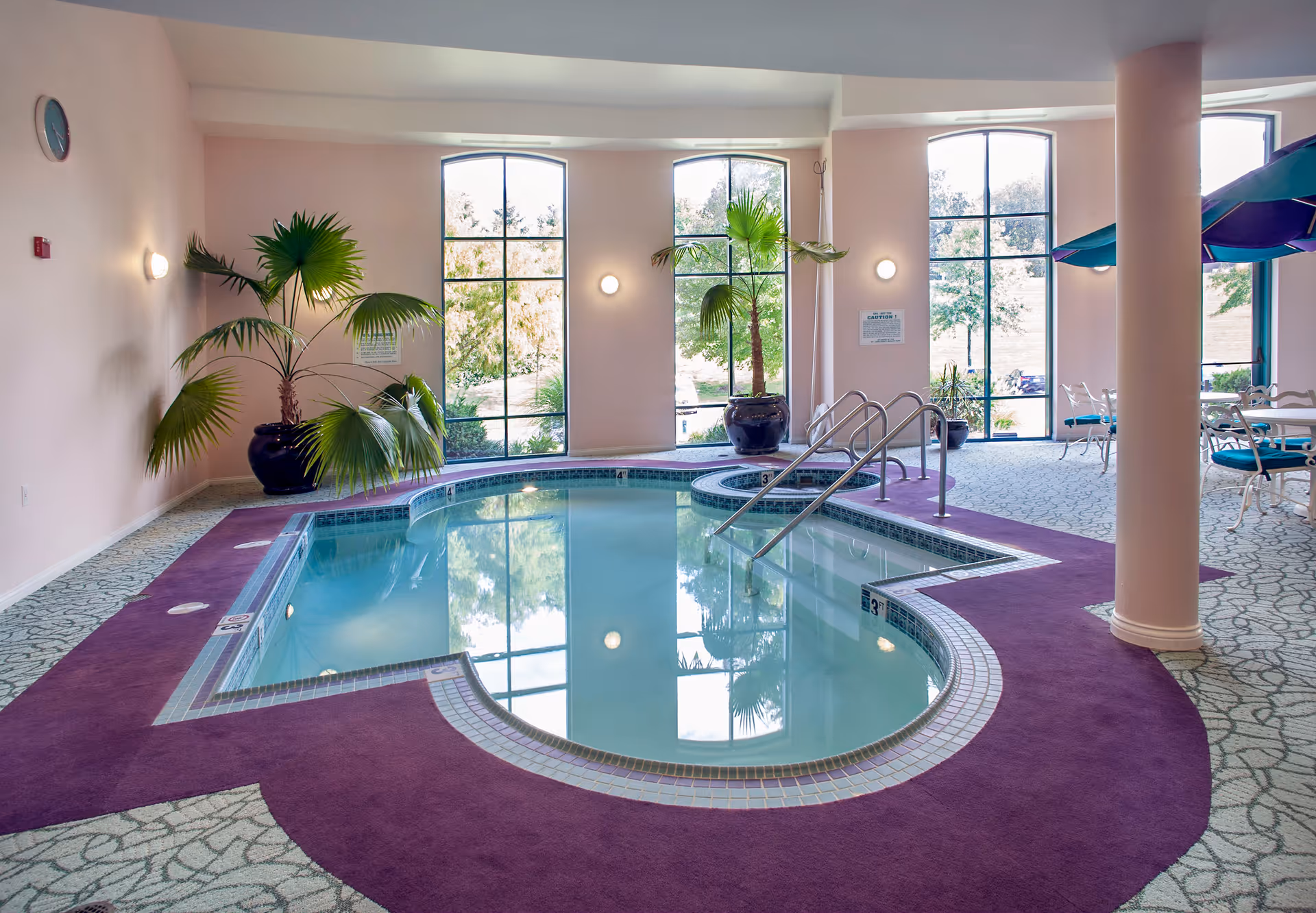 Indoor pool area with a uniquely shaped pool surrounded by purple carpeting and patterned flooring. Large windows let in natural light and show outdoor greenery. There are potted palm plants near the windows and a seating area with tables and chairs under umbrellas to the right.