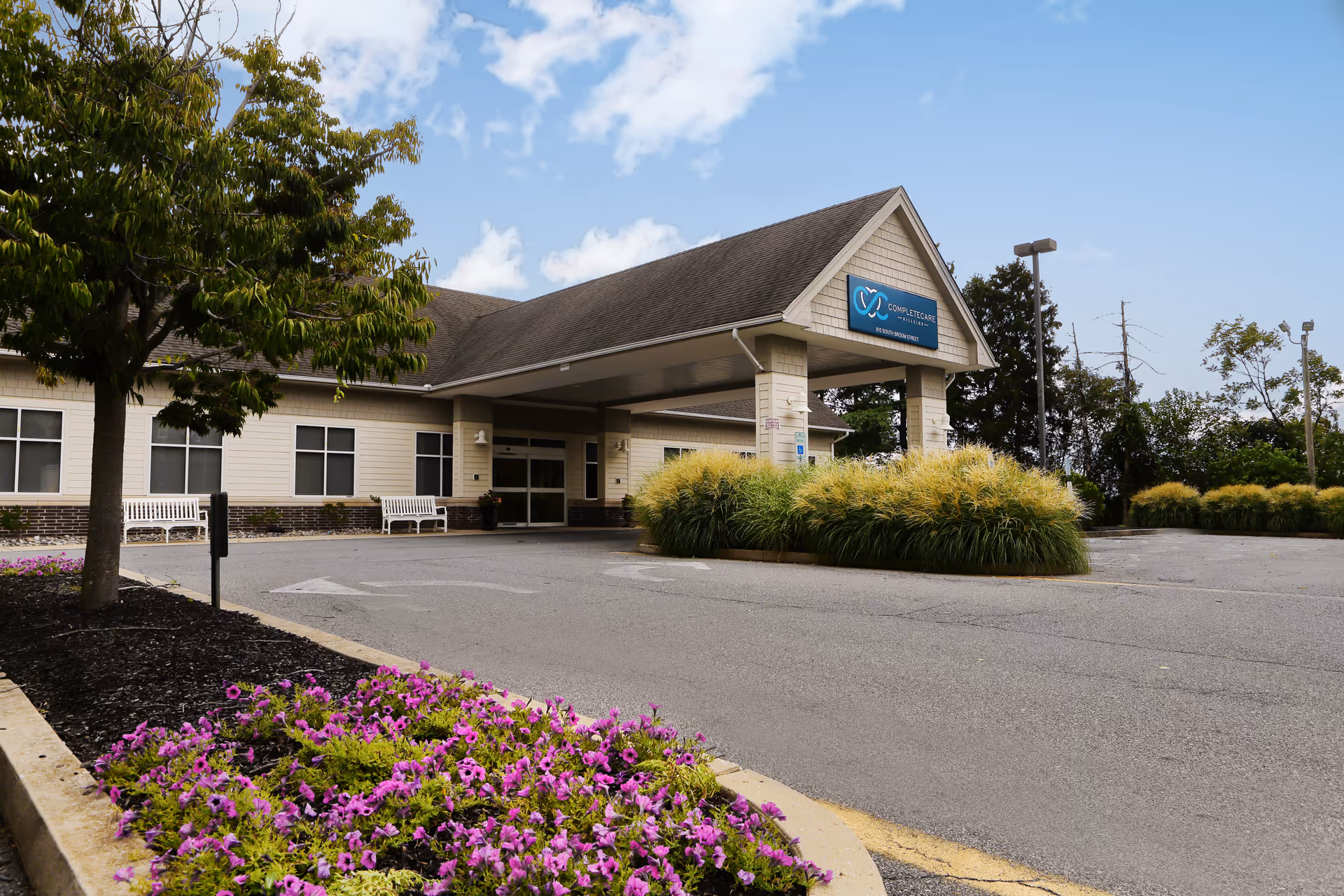 Exterior view of Complete Care at Hillside facility showing the main entrance with a covered drop-off area, surrounded by landscaped greenery including bushes and purple flowers, with a tree on the left and benches near the building.