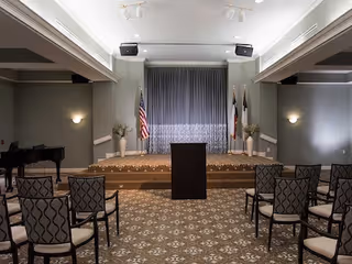 An event room with a small stage at the front featuring a podium, two American flags, and two other flags. The room has rows of chairs with patterned upholstery facing the stage. The walls are painted gray with wall sconces providing light, and there is a grand piano on the left side of the room.