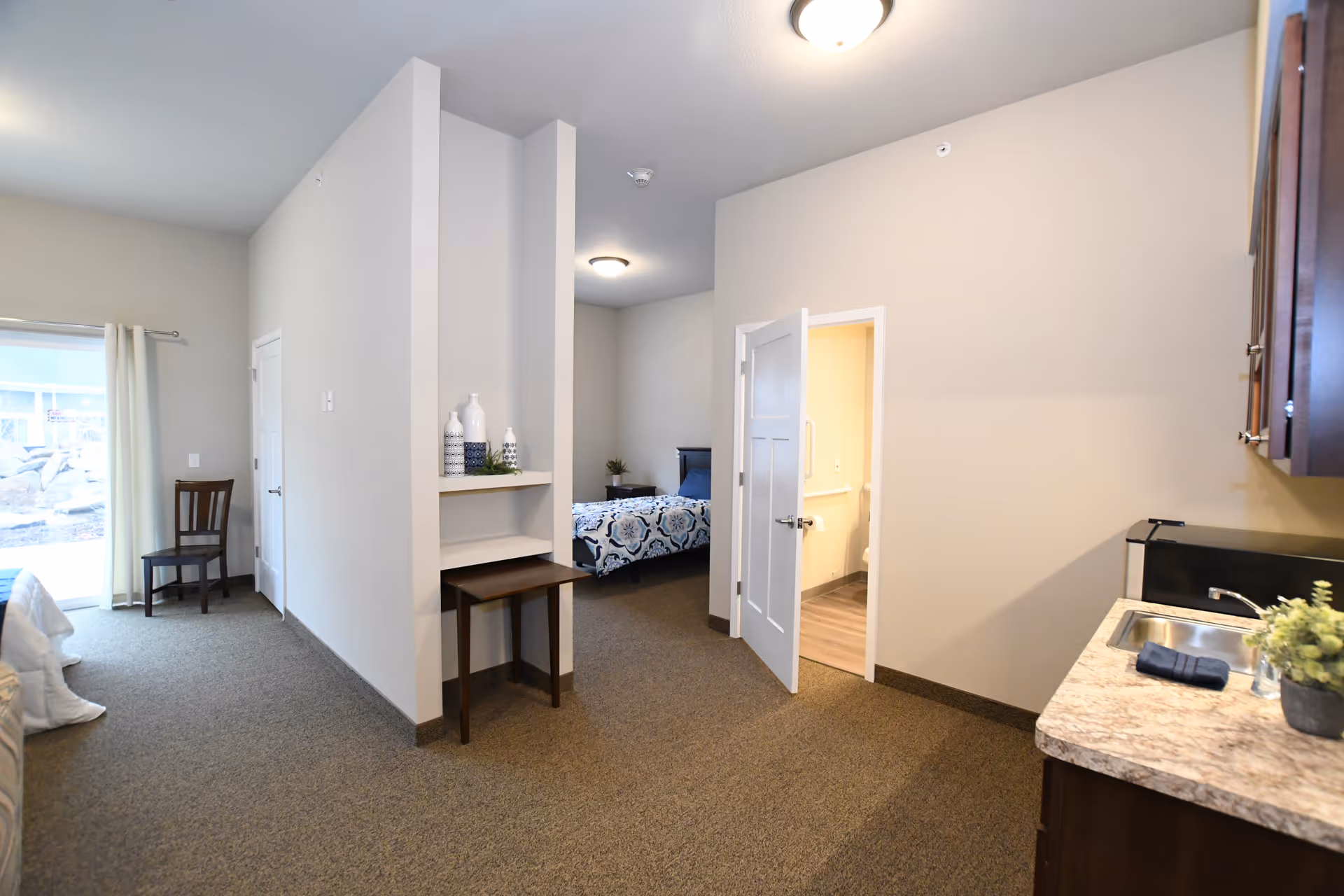 Interior view of a senior living facility room showing a small kitchenette with a sink and countertop on the right, a partially open door leading to a bathroom, a bedroom area with a bed and nightstand in the background, and a chair near a sliding glass door with curtains on the left.
