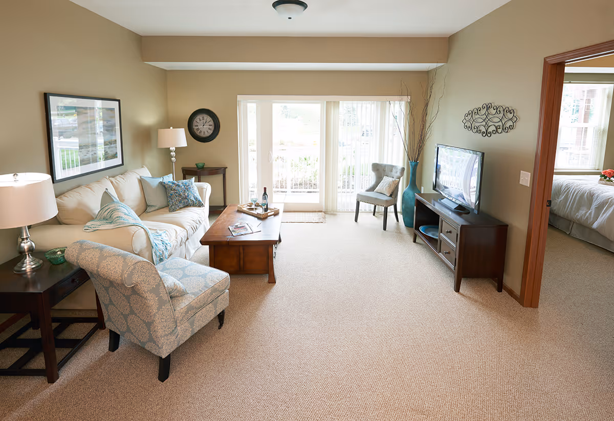A bright and cozy living room with beige walls and carpet. The room features a cream-colored sofa with blue and patterned throw pillows, a wooden coffee table with a tray holding a bottle of wine and glasses, a patterned armchair, and a side table with a lamp. A flat-screen TV sits on a wooden TV stand against the right wall, next to a large blue vase with decorative branches. Sliding glass doors with vertical blinds lead to an outdoor area. A doorway on the right reveals a bedroom with a bed and window.