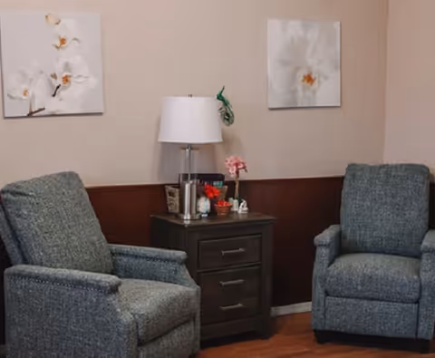 Two gray upholstered armchairs positioned on either side of a dark wooden side table with three drawers. On the table is a white lamp, a small basket, and decorative items including a small pink flower arrangement. The wall behind features two framed pictures of white orchids.