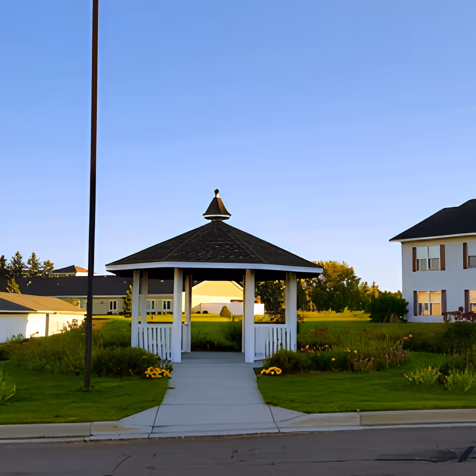A small white gazebo with a dark shingled roof situated on a concrete pathway surrounded by green grass and flower beds, with buildings and trees in the background under a clear blue sky.