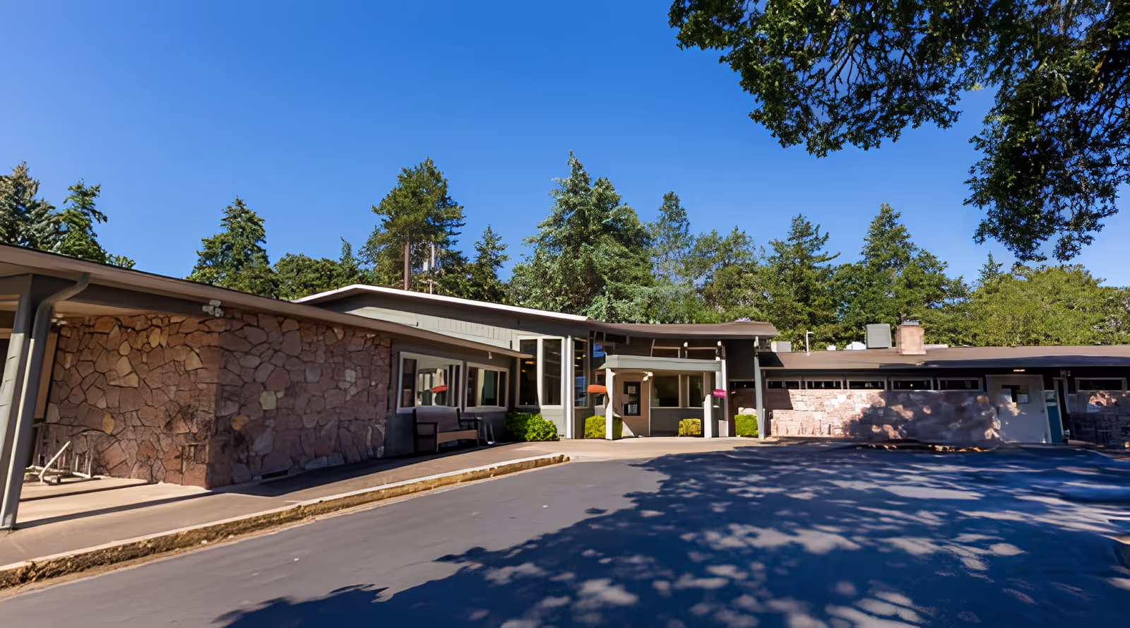 Front exterior of a single-story rehabilitation center with stone facades, entrance canopy, driveway, and surrounding trees under a clear blue sky.